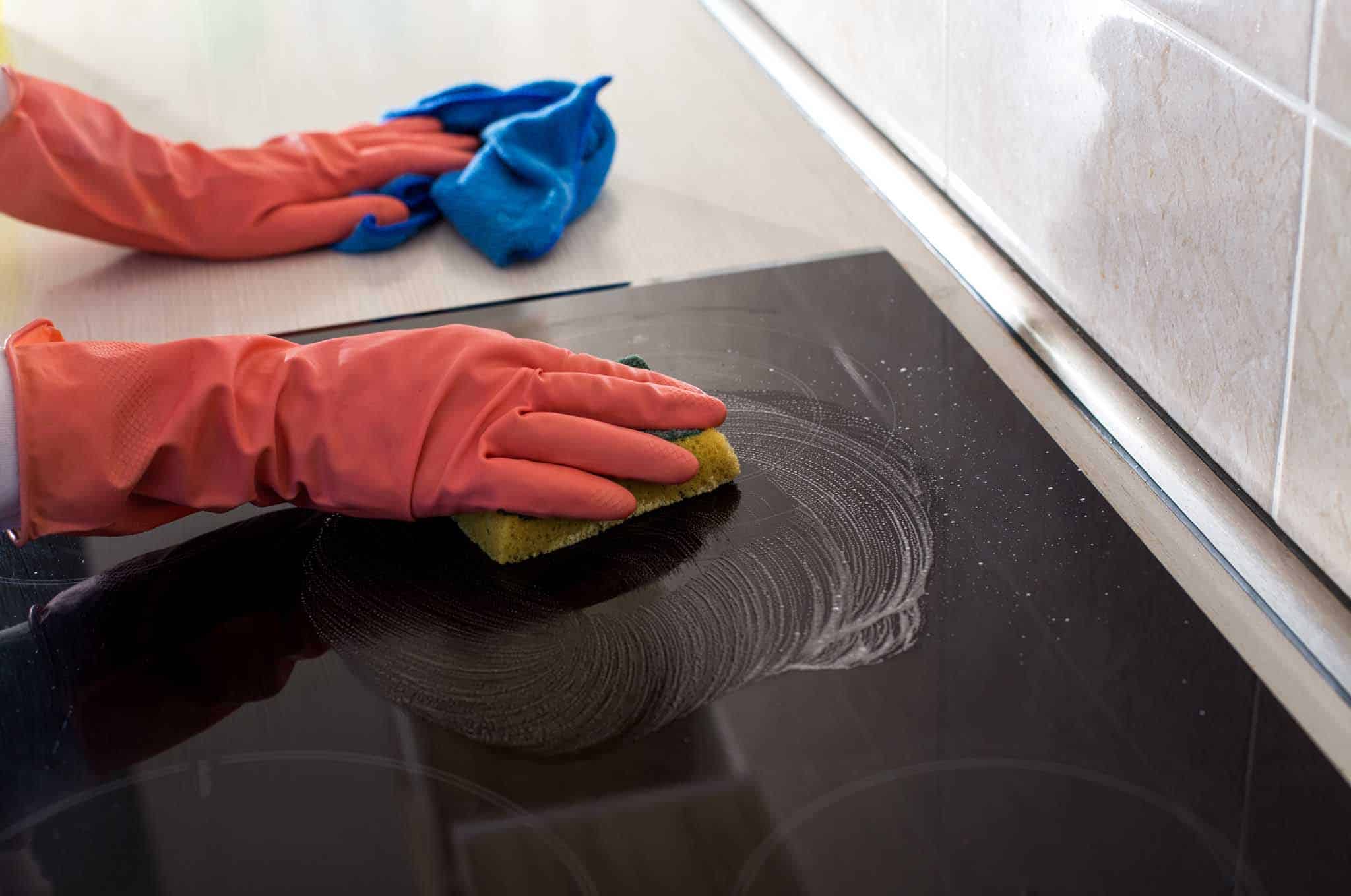 En person med rosa gummihansker bruker en gul svamp og en blå klut til å vaske en sort, keramisk koketopp på et kjøkken.